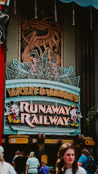 A theme park attraction entrance featuring a vibrant sign with characters and colorful lights. The sign reads 'Mickey & Minnie's Runaway Railway' and there are people walking beneath it. Above the sign, an intricate dragon design is incorporated into the structure.