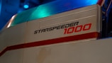 A close-up of a futuristic vehicle with the label 'Starspeeder 1000' prominently displayed in bold, stylized text. The design is sleek and angular, featuring a mix of metallic and colorful surfaces. The environment suggests an industrial or sci-fi setting with a dark, glowing blue light reflecting off parts of the vehicle.
