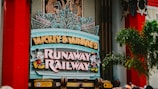 A colorful sign featuring playful typography and animated figures associated with a popular entertainment franchise. The sign is adorned with neon lights and decorative elements, set against a backdrop of bright red pillars and greenery.