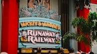 A colorful sign featuring playful typography and animated figures associated with a popular entertainment franchise. The sign is adorned with neon lights and decorative elements, set against a backdrop of bright red pillars and greenery.