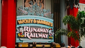 A colorful sign featuring playful typography and animated figures associated with a popular entertainment franchise. The sign is adorned with neon lights and decorative elements, set against a backdrop of bright red pillars and greenery.