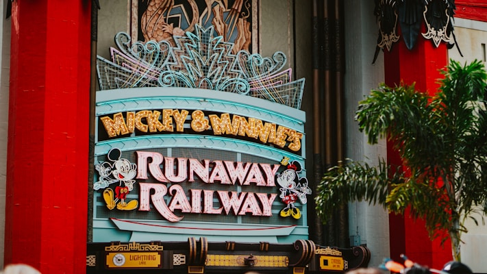 A colorful sign featuring playful typography and animated figures associated with a popular entertainment franchise. The sign is adorned with neon lights and decorative elements, set against a backdrop of bright red pillars and greenery.