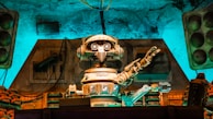 A vintage-style robot with a humanoid form, featuring round eyes and detailed mechanical arms, is positioned in a technology-filled environment. The setting has an old-fashioned industrial vibe, with a mixture of mechanical equipment and stacks of components seen behind the robot. There is a notable use of ambient turquoise and brown lighting that highlights the metallic textures and creates a retro atmosphere.