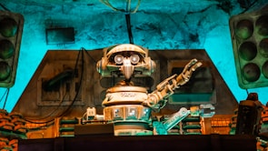 A vintage-style robot with a humanoid form, featuring round eyes and detailed mechanical arms, is positioned in a technology-filled environment. The setting has an old-fashioned industrial vibe, with a mixture of mechanical equipment and stacks of components seen behind the robot. There is a notable use of ambient turquoise and brown lighting that highlights the metallic textures and creates a retro atmosphere.
