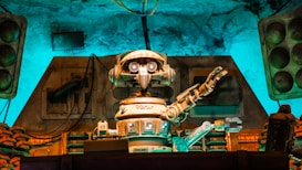 A vintage-style robot with a humanoid form, featuring round eyes and detailed mechanical arms, is positioned in a technology-filled environment. The setting has an old-fashioned industrial vibe, with a mixture of mechanical equipment and stacks of components seen behind the robot. There is a notable use of ambient turquoise and brown lighting that highlights the metallic textures and creates a retro atmosphere.