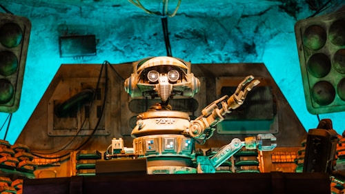 A vintage-style robot with a humanoid form, featuring round eyes and detailed mechanical arms, is positioned in a technology-filled environment. The setting has an old-fashioned industrial vibe, with a mixture of mechanical equipment and stacks of components seen behind the robot. There is a notable use of ambient turquoise and brown lighting that highlights the metallic textures and creates a retro atmosphere.