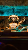 A retro-styled robot with large eyes and a mechanical arm extending outward is the focal point. The scene features a background with various industrial elements including wires and pipes, all bathed in a blue and orange light.