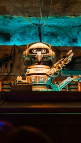 A retro-styled robot with large eyes and a mechanical arm extending outward is the focal point. The scene features a background with various industrial elements including wires and pipes, all bathed in a blue and orange light.