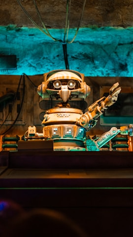 A retro-styled robot with large eyes and a mechanical arm extending outward is the focal point. The scene features a background with various industrial elements including wires and pipes, all bathed in a blue and orange light.