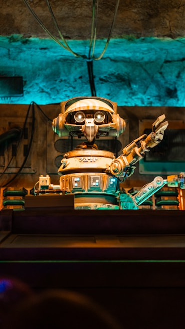A retro-styled robot with large eyes and a mechanical arm extending outward is the focal point. The scene features a background with various industrial elements including wires and pipes, all bathed in a blue and orange light.