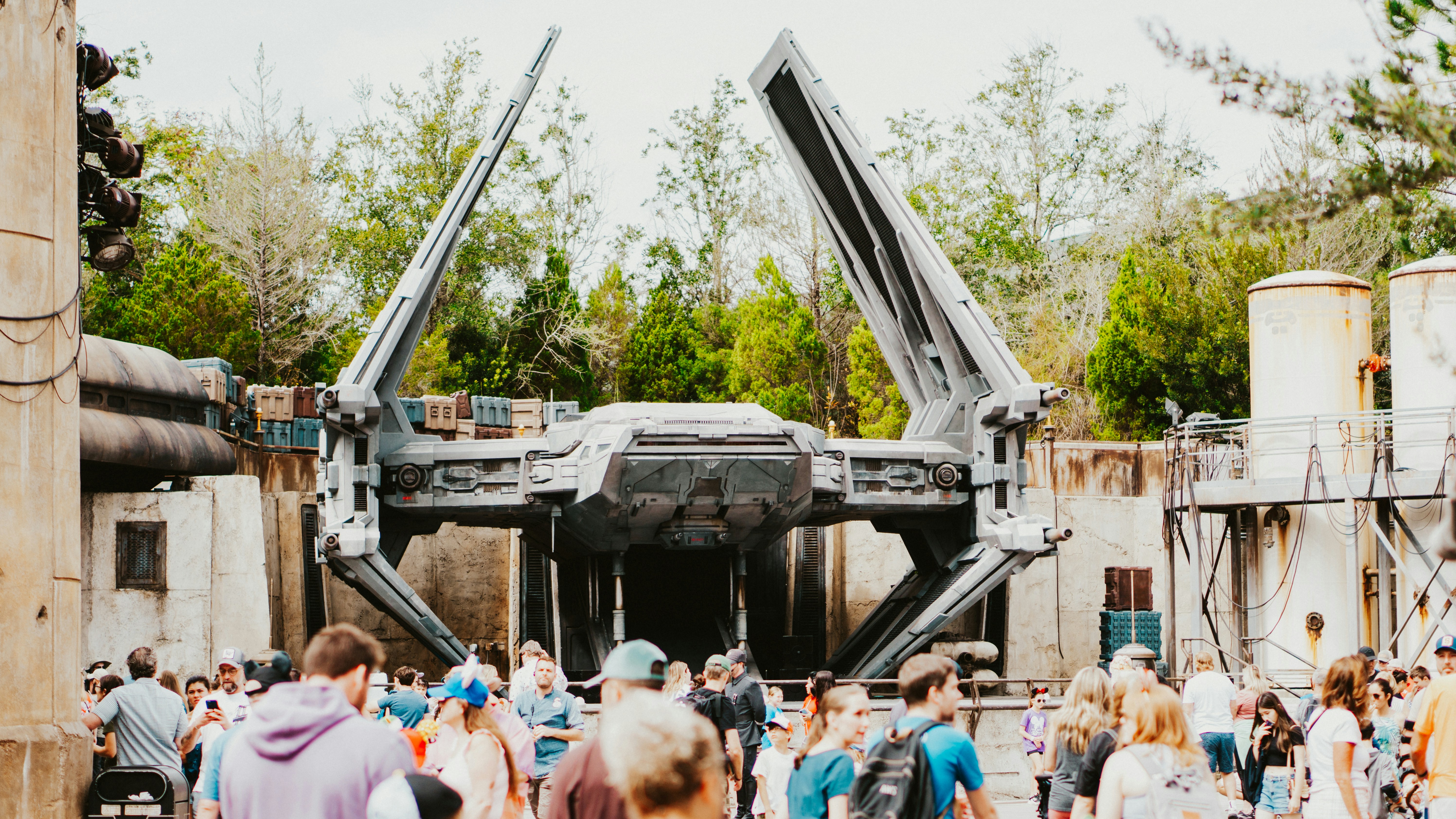 A large spacecraft with extended wings is displayed in an outdoor setting. The scene is lively, with a crowd of people gathered in the foreground. The background includes trees and industrial-looking structures, suggesting a themed park or exhibition area.