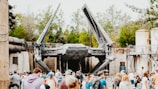 A large spacecraft with extended wings is displayed in an outdoor setting. The scene is lively, with a crowd of people gathered in the foreground. The background includes trees and industrial-looking structures, suggesting a themed park or exhibition area.