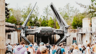 A large spacecraft with extended wings is displayed in an outdoor setting. The scene is lively, with a crowd of people gathered in the foreground. The background includes trees and industrial-looking structures, suggesting a themed park or exhibition area.