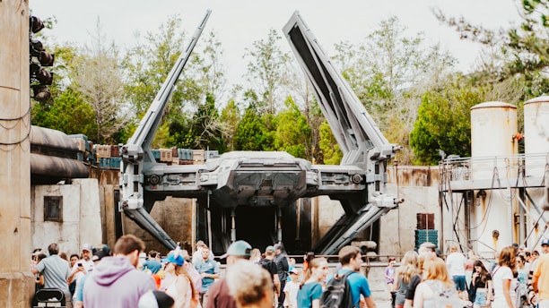 A large spacecraft with extended wings is displayed in an outdoor setting. The scene is lively, with a crowd of people gathered in the foreground. The background includes trees and industrial-looking structures, suggesting a themed park or exhibition area.