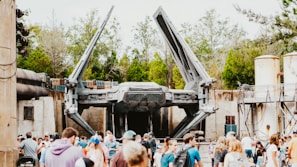 A large spacecraft with extended wings is displayed in an outdoor setting. The scene is lively, with a crowd of people gathered in the foreground. The background includes trees and industrial-looking structures, suggesting a themed park or exhibition area.