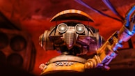 A robot with large circular eyes, a mechanical mouthpiece, and an articulated arm set against a warm, reddish background. The metal features have a worn, rustic appearance, adding a sense of vintage charm.