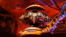 A robot with large circular eyes, a mechanical mouthpiece, and an articulated arm set against a warm, reddish background. The metal features have a worn, rustic appearance, adding a sense of vintage charm.