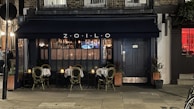 A cozy outdoor seating area of a small restaurant with a dark navy blue awning displaying the name 'ZOILO'. The tables are set with white tablecloths and candles, creating an inviting and intimate atmosphere. The warm glow of lights inside the restaurant spills out through large windows.