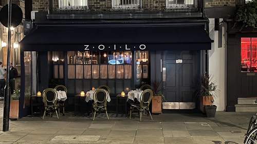 A cozy outdoor seating area of a small restaurant with a dark navy blue awning displaying the name 'ZOILO'. The tables are set with white tablecloths and candles, creating an inviting and intimate atmosphere. The warm glow of lights inside the restaurant spills out through large windows.