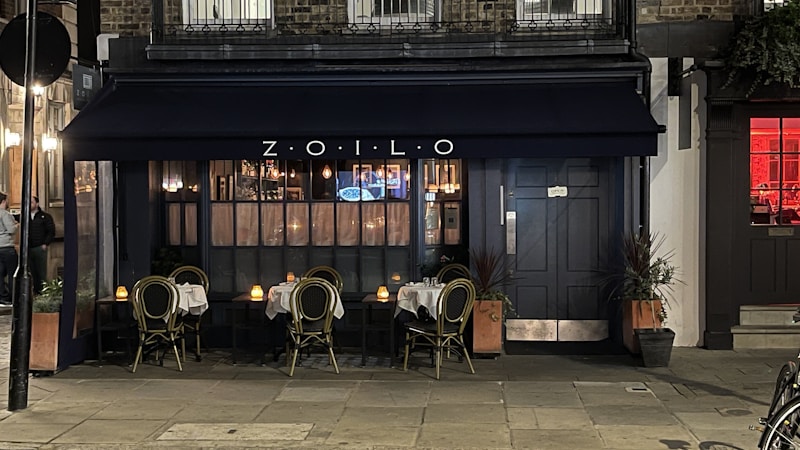 A cozy outdoor seating area of a small restaurant with a dark navy blue awning displaying the name 'ZOILO'. The tables are set with white tablecloths and candles, creating an inviting and intimate atmosphere. The warm glow of lights inside the restaurant spills out through large windows.