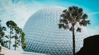 A large geodesic sphere with a faceted surface stands prominently in a landscaped park setting. Several trees, including a palm tree, are positioned in the foreground with blue sky visible above.
