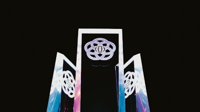 Three tall, reflective structures with geometric designs rise against a dark background. The structures display interconnected circular patterns, with a globe symbol at their center.