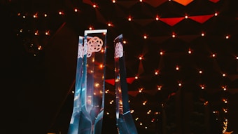 Tall, transparent, geometric sculptures stand illuminated against a dark, textured background. The structures have intricate designs near the top, while warm, reddish lighting creates a dramatic ambiance.