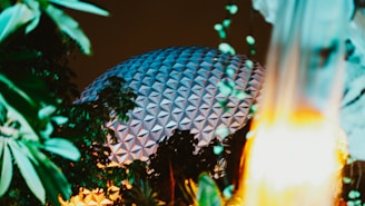 A geometric dome-like structure is partially obscured by lush green foliage and illuminated by warm, ambient lighting against a dark sky. The scene captures a mix of architectural and natural elements, creating a harmonious blend of nature and modern design.