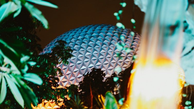 A geometric dome-like structure is partially obscured by lush green foliage and illuminated by warm, ambient lighting against a dark sky. The scene captures a mix of architectural and natural elements, creating a harmonious blend of nature and modern design.