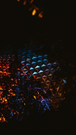 A nighttime scene featuring a geodesic dome partially obscured by silhouetted foliage. The dome is adorned with a geometric pattern and illuminated by vibrant colored lights in shades of blue, orange, and red, creating a striking visual contrast against the dark sky.