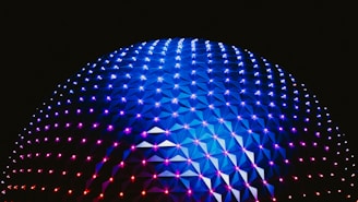A large geodesic dome illuminated with vibrant blue, purple, and pink lights against a dark background. The interconnected triangular panels create a striking pattern that enhances the futuristic aesthetic of the structure.