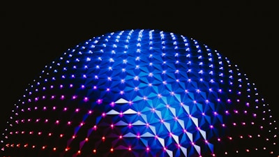 A large geodesic dome illuminated with vibrant blue, purple, and pink lights against a dark background. The interconnected triangular panels create a striking pattern that enhances the futuristic aesthetic of the structure.