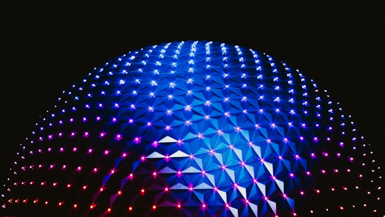 A large geodesic dome illuminated with vibrant blue, purple, and pink lights against a dark background. The interconnected triangular panels create a striking pattern that enhances the futuristic aesthetic of the structure.