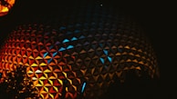 A geodesic dome with a reflective, triangular-patterned surface is illuminated by vibrant red, orange, and blue lights shining against the dark night sky. Silhouettes of trees are visible in the foreground.