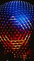 An illuminated geometric sphere with a pattern of interconnected triangles and vibrant lights. The top half of the structure is lit with shades of blue and purple, while the bottom half transitions into red and orange hues.