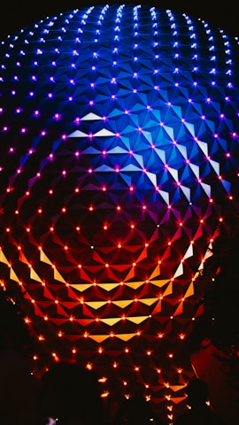 An illuminated geometric sphere with a pattern of interconnected triangles and vibrant lights. The top half of the structure is lit with shades of blue and purple, while the bottom half transitions into red and orange hues.