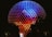 A large geodesic sphere is illuminated with vibrant colors in a gradient pattern from blue at the top to red at the bottom. The structure is covered in triangular panels that reflect the light, creating a dazzling effect against the night sky. Small lights highlight the edges, enhancing the geometric appearance.