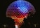 A large geodesic sphere is illuminated with vibrant colors in a gradient pattern from blue at the top to red at the bottom. The structure is covered in triangular panels that reflect the light, creating a dazzling effect against the night sky. Small lights highlight the edges, enhancing the geometric appearance.