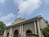 A neoclassical building with tall columns and arched windows, featuring traditional architectural elements. The structure is topped with a flag showing the red national flag with yellow stars. The sky is clear with a few clouds, and there is greenery surrounding the building.
