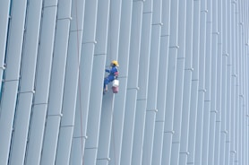 A person wearing safety gear is suspended by ropes, performing maintenance or cleaning on the side of a large building with vertical panels.