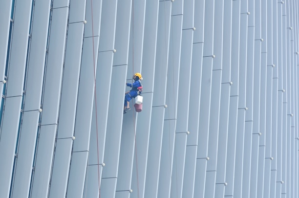 A person wearing safety gear is suspended by ropes, performing maintenance or cleaning on the side of a large building with vertical panels.