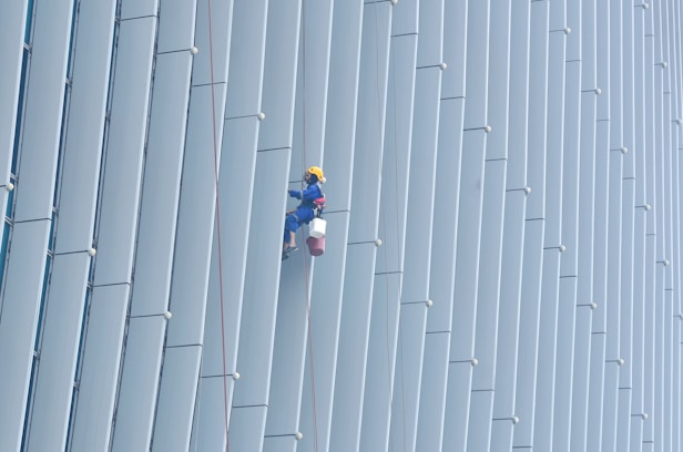 A person wearing safety gear is suspended by ropes, performing maintenance  cleaning on the side of a large building with vertical panels.