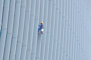 A person wearing safety gear is suspended by ropes, performing maintenance or cleaning on the side of a large building with vertical panels.