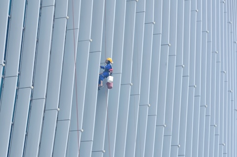 A person wearing safety gear is suspended by ropes, performing maintenance or cleaning on the side of a large building with vertical panels.