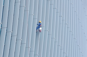 A person wearing safety gear is suspended by ropes, performing maintenance or cleaning on the side of a large building with vertical panels.