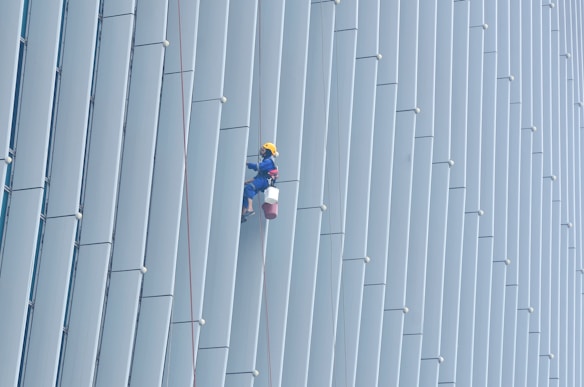 A person wearing safety gear is suspended by ropes, performing maintenance or cleaning on the side of a large building with vertical panels.