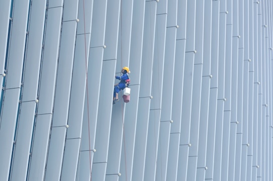 A person wearing safety gear is suspended by ropes, performing maintenance or cleaning on the side of a large building with vertical panels.
