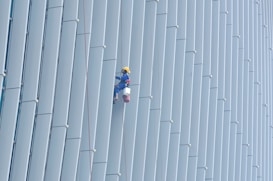 A person wearing safety gear is suspended by ropes, performing maintenance or cleaning on the side of a large building with vertical panels.