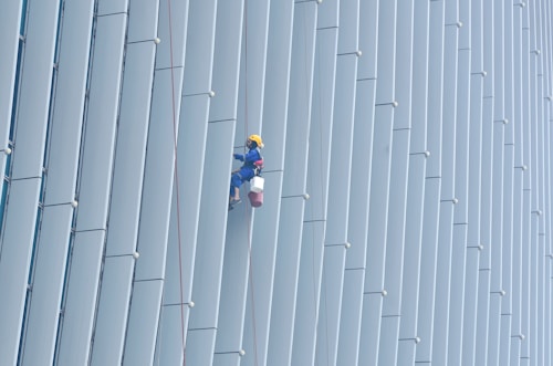 A person wearing safety gear is suspended by ropes, performing maintenance or cleaning on the side of a large building with vertical panels.