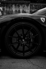 A black and white photograph focusing on the front wheel of a sports car. The car has a sleek body with a prominent badge on the side panel. The tire and hubcap are detailed, showcasing a stylish design with visible brake components. A person is faintly visible in the background standing near a wall.